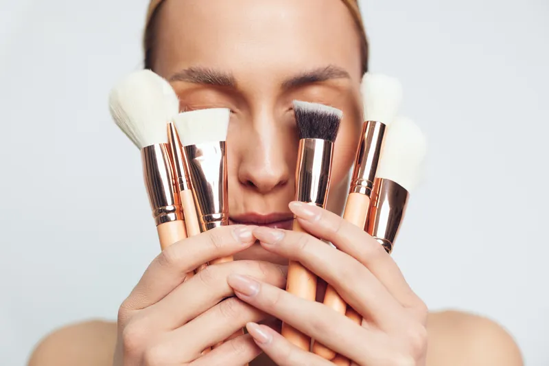 A woman holds various makeup brushes in front of her face with closed eyes. The image focuses on beauty tools used for applying cosmetics on a plain background.