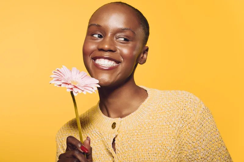 Smiling black woman with dark skin holds pink flower against yellow background wearing yellow textured sweater. Concept of happiness, beauty, natural, joy, and floral positive mood.