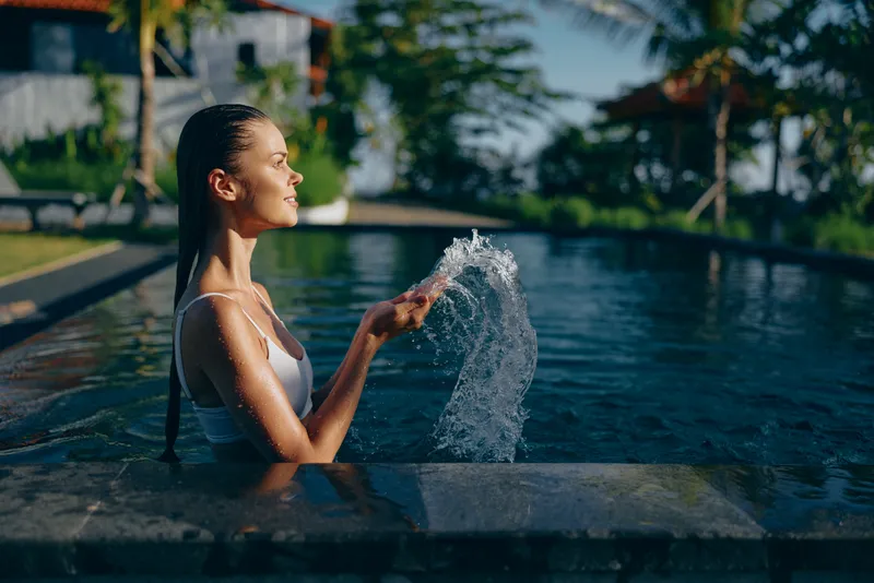 Relaxed woman in a pool splashes a fountain stream, enjoying a sunny outdoor moment. Tropical garden backdrop, clear blue water, serene mood, wellness lifestyle, self care, calm atmosphere, leisure.