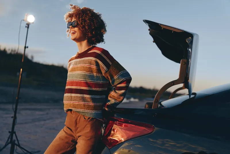 Cheerful woman lifestyle outdoors by open car trunk at sunset, curly hair glowing in warm light, relaxed smile, striped sweater and autumn jacket, road background, evening travel vibe