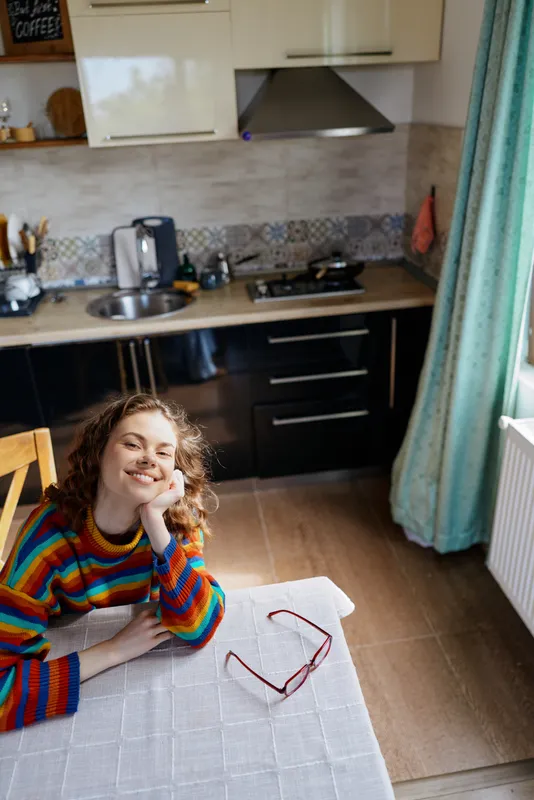 Cheerful woman indoors at a kitchen table, wearing a colorful sweater, relaxed pose, warm light, candid lifestyle moment, casual home setting, friendly mood and soft ambience