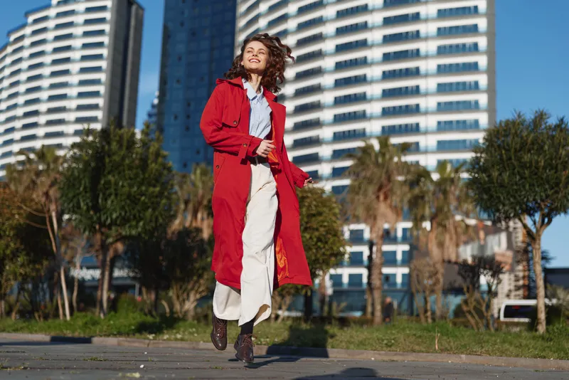 Young woman in a red coat walks along a city street with tall buildings behind, wearing white trousers and black boots, enjoying a bright urban day.