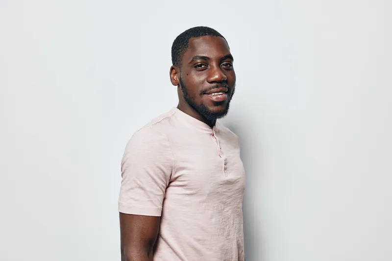 African American man with dark skin and beard smiling in casual light pink shirt standing against white background in studio portrait with confident expression.