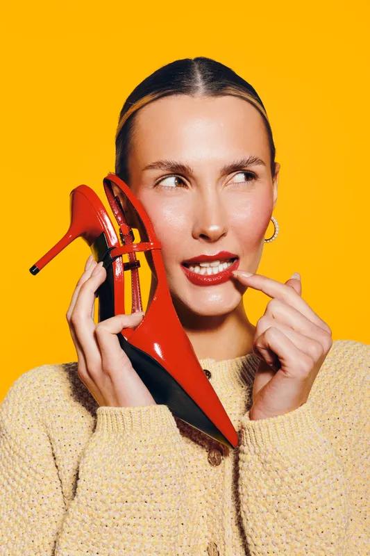Young woman in beige sweater holds red high heel shoe near face, looks sideways with thoughtful expression on bright yellow background. Concept of fashion, style, and creativity.
