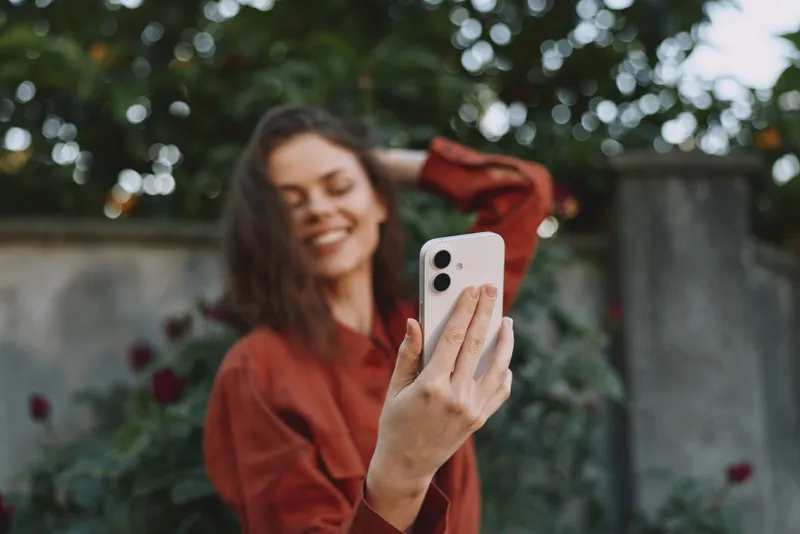 Woman lifestyle selfie outdoors in a casual fashion jacket, smiling while holding smartphone for a portrait shot in a sunlit garden, relaxed mood. Bright background with greenery adds cheer