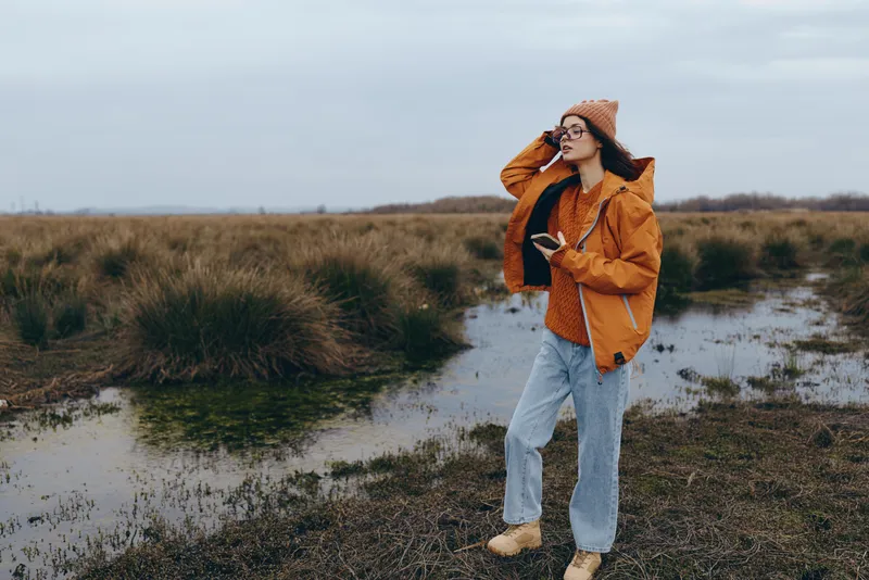 Woman lifestyle outdoors in autumn landscape, walking along a narrow muddy path beside a grassy field, wearing orange jacket and straw hat, adjusting hair, cloudy sky, natural tones