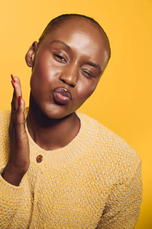 Close-up of a dark skinned African woman with short hair wearing a yellow sweater, making a playful kiss face and winking against a bright yellow background, concept of joy and fun.