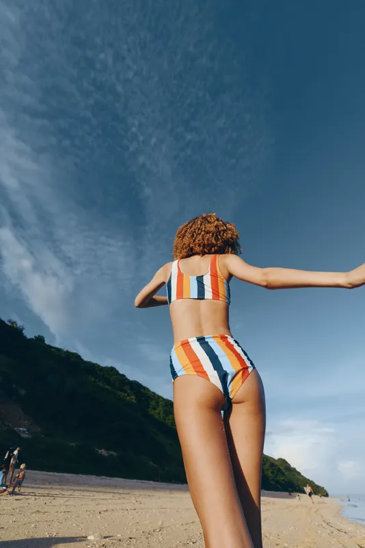 Young woman in striped swimsuit seen from back view walks along beach with arms out, enjoying summer vacation by the ocean, sunny sky, sand and gentle waves, outdoor leisure scene in peace