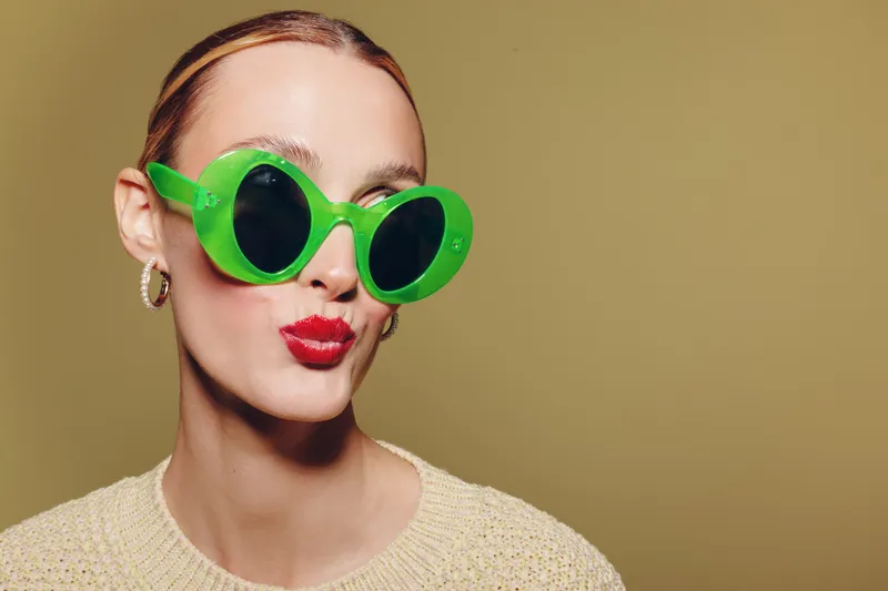 Young woman with green round sunglasses and red lipstick makes a playful kiss face. She wears a beige sweater and hoop earrings on a plain olive background. Concept of fashion and fun.