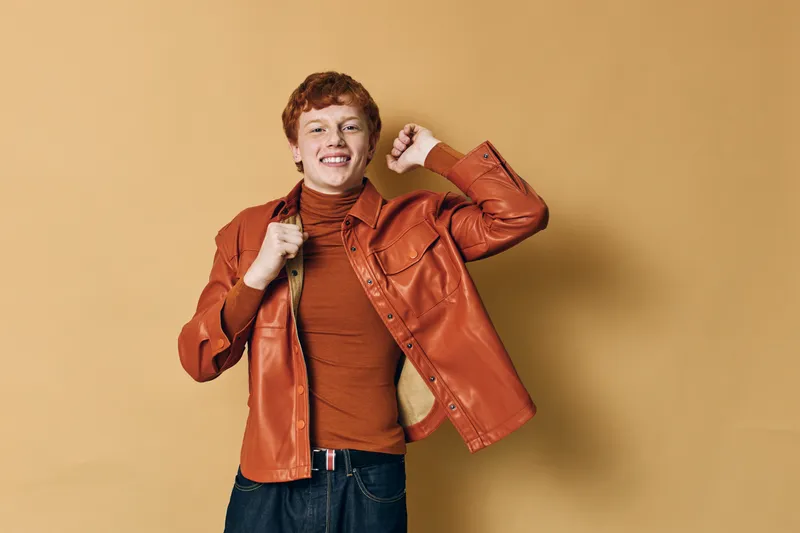 Young man with short red hair wearing orange leather jacket and turtleneck smiling with raised fist for celebration and confidence on neutral background studio.