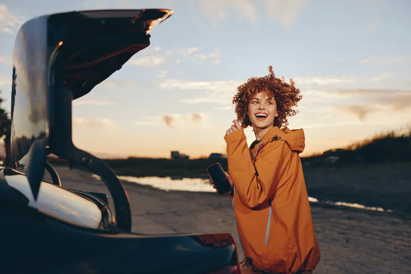 Lifestyle woman in orange jacket stands beside open car at beach at sunset, with a smile and talking on a phone, warm golden light, travel moment, outdoors scene, for vacation right now