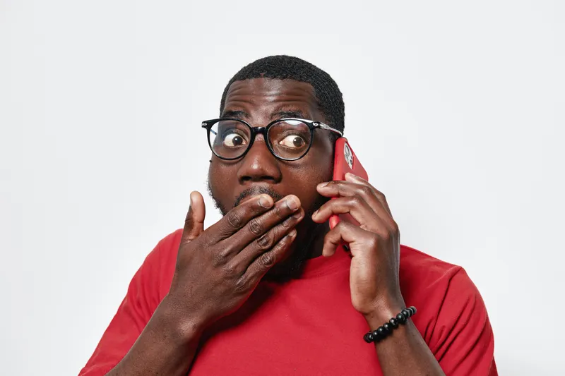 Black man with dark skin wearing glasses and red shirt talking on phone with surprised expression covering mouth with hand on white background.