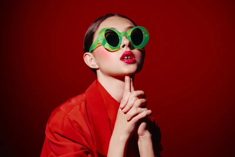 Stylish woman in bright red jacket and bold green retro sunglasses poses thoughtfully against an intense red backdrop. Perfect for beauty creative fashion concepts.