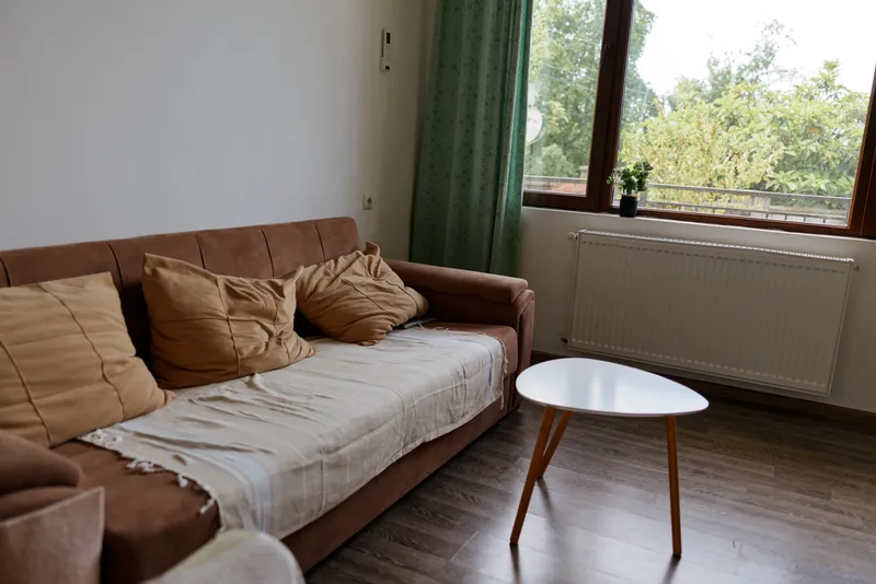 Bright living room scene with beige sofa and plush cushions, minimal decor, large window letting warm sunlight on wooden floor, cozy interior for home living