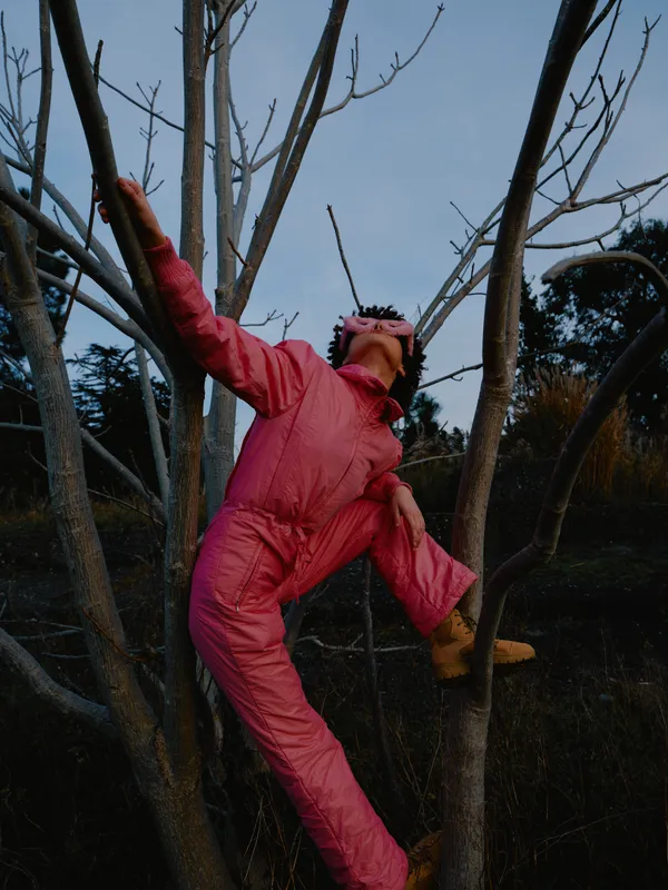 Person in pink snowsuit climbs a tree balancing on branches during dusk outdoor adventure playful exploration in a winter forest energetic pose and vivid clothing in a natural setting