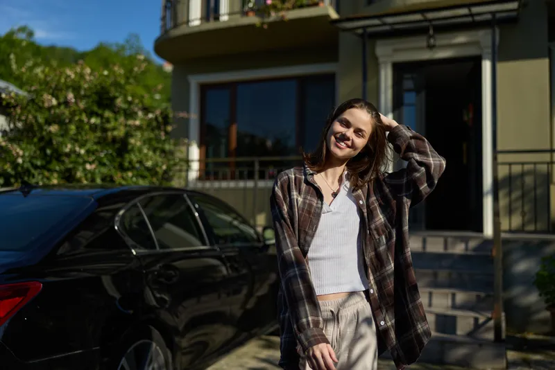 A young woman in a plaid shirt and white top stands beside a dark car on a sunny street, smiling and relaxed, capturing a casual lifestyle moment outdoors