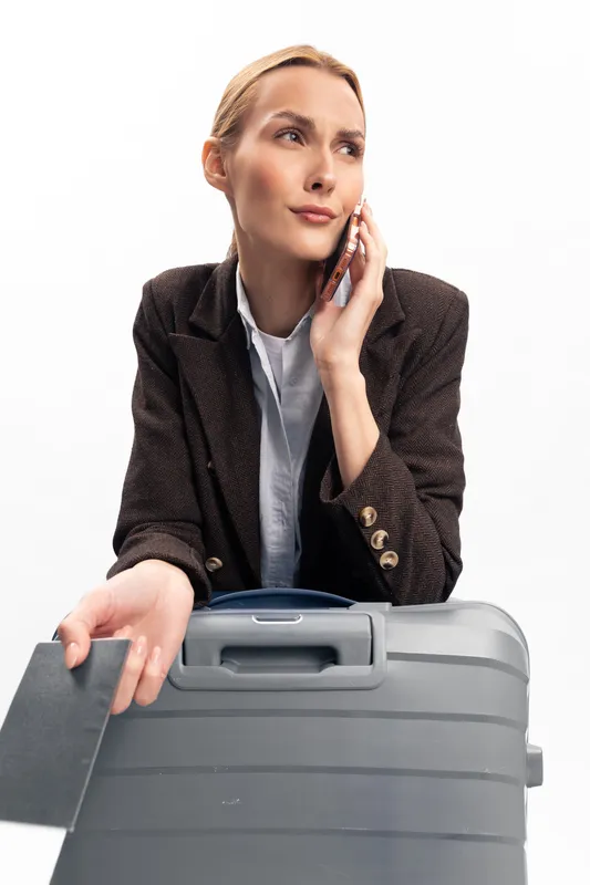 Young woman in dark blazer talking on phone while holding passport and suitcase. Concept of travel, business trip, communication, and female traveler with luggage on white background.