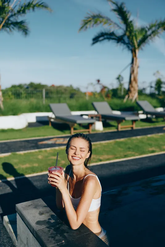 Young woman in a white bikini enjoying a refreshing drink while relaxing in an outdoor pool on a sunny day. Palm trees and lounge chairs create a tropical lifestyle atmosphere.