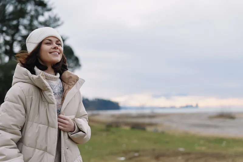 Woman in a warm coat and beanie stands outdoors along the coast, smiling softly. A calm sea and muted sky create a tranquil, chilly mood around her peaceful pose.
