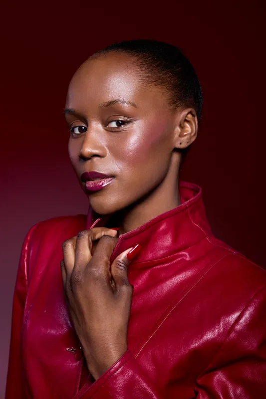 Portrait of confident African woman with dark skin wearing red leather jacket, looking at camera with subtle smile. Studio lighting highlights smooth glowing skin on red background.