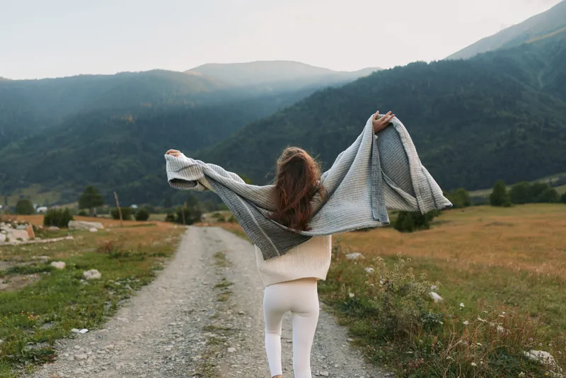 A young woman walks along a rural path through a sunlit valley, arms raised as a knitted shawl billows behind her, expressing freedom and outdoor lifestyle.