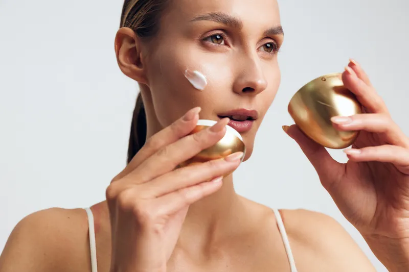 Young woman applying white face cream on cheek holding golden jar close to face. Skincare routine concept with natural makeup and soft lighting on white background.