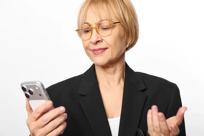 Senior woman with glasses, holding a smartphone and gesturing with hands, smiling in a business suit, indoor portrait on a clean white background, studio light
