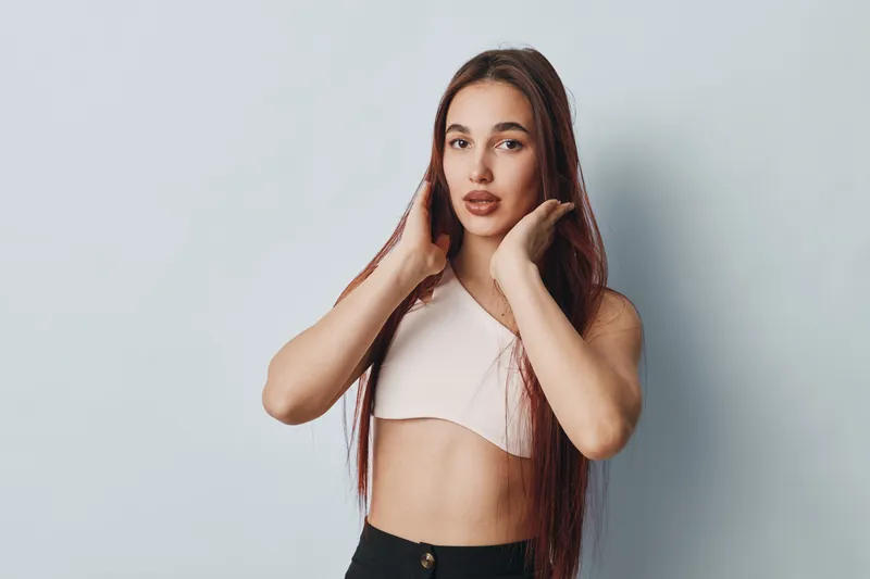 Young woman with long hair posing in light top and black pants against plain background. Female model with natural makeup and confident expression for fashion or beauty concept.