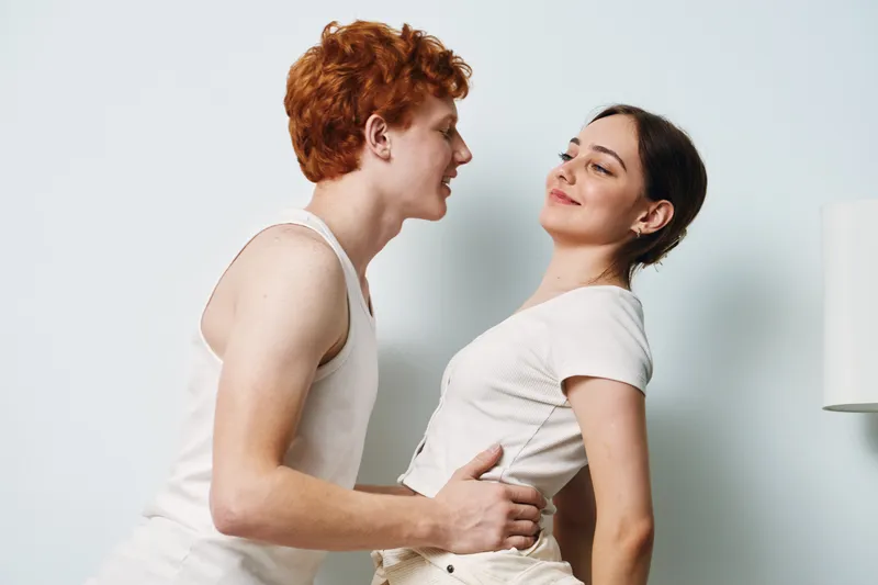 Young man and woman in casual outfits embracing with playful and affectionate expressions in a bright room. Couple interaction with smiling, care, and connection.
