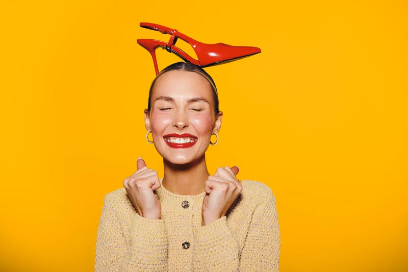 Happy woman with red lipstick and earrings smiles with closed eyes, holding red high heel shoes on her head against bright yellow background. Concept of joy and fashion fun.