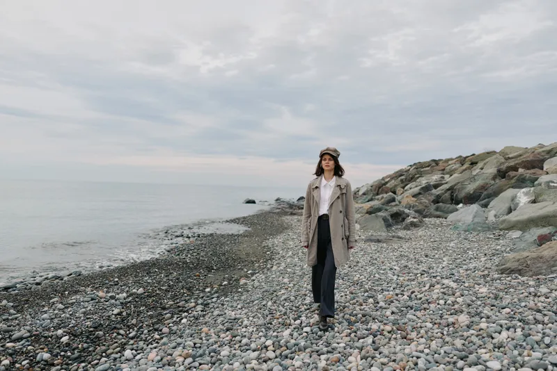 Young woman in coat and hat walks alone on rocky beach under cloudy sky. Calm sea and rugged shore create moody natural landscape. Concept of solitude, autumn, and nature walk.