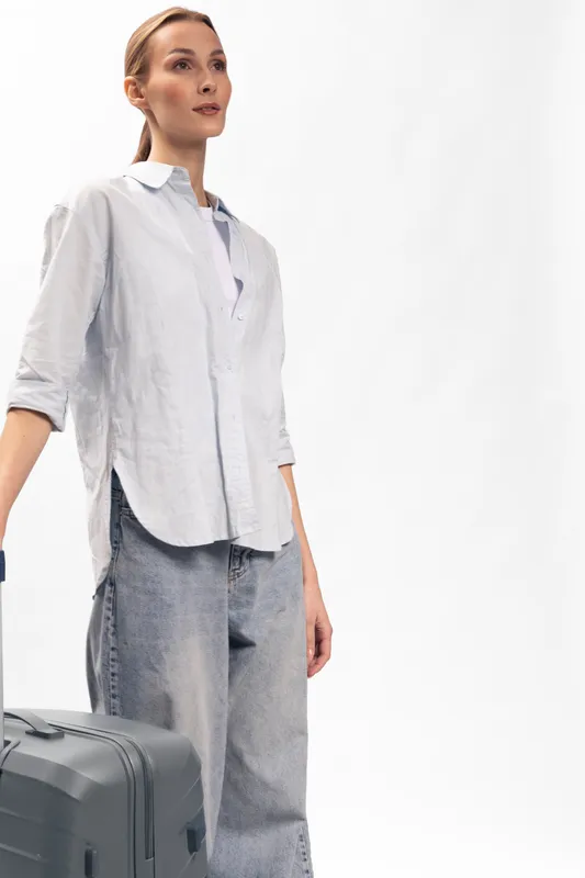 Young woman in casual shirt and jeans stands with suitcase on white background. Travel concept with luggage and modern style. Minimalist studio photo with natural light.