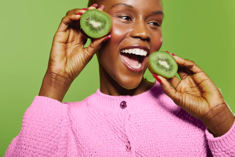 Happy African woman with dark skin holds two kiwi halves near face smiling wide on green background. Concept of healthy lifestyle, fresh fruit, nutrition, and joyful mood.