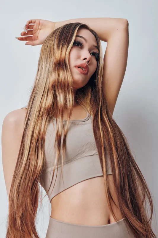 Young woman with long hair posing in beige sportswear top standing against plain background with arm raised above head for fashion portrait and fitness style photo.