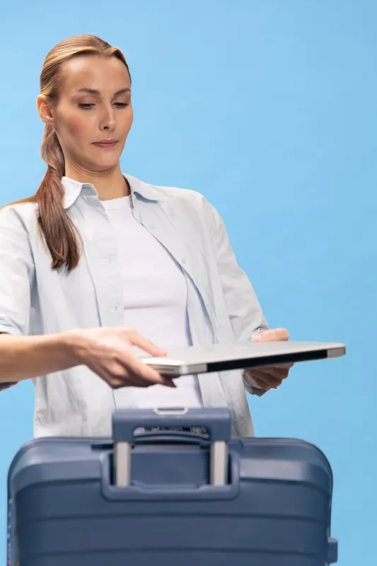 Young woman holding laptop over suitcase preparing for travel, wearing casual white shirt. Concept of business trip, packing, and travel planning on blue background.