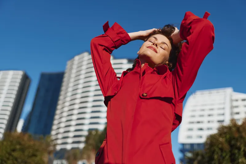 Young woman in a red jacket enjoys fresh air with closed eyes and raised arms against modern city buildings on a sunny day.