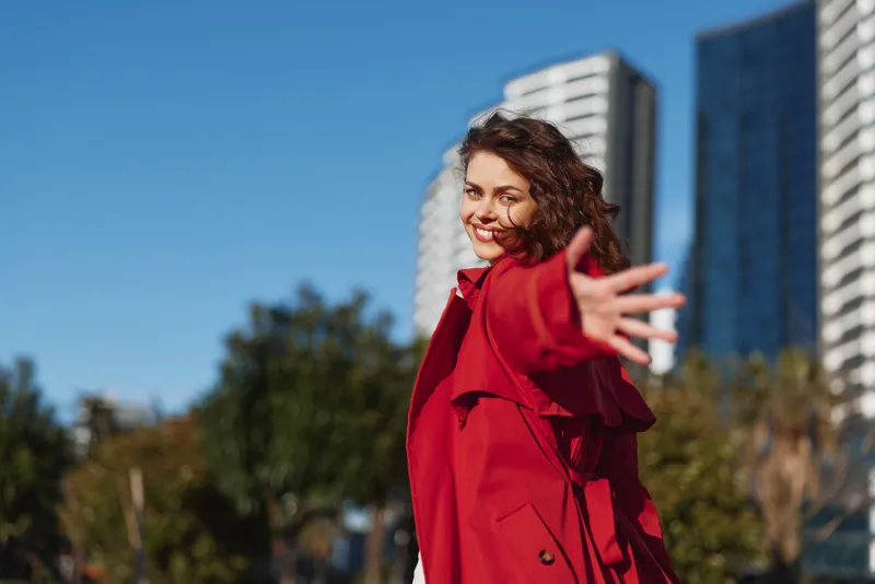 Young woman with curly hair wearing a red coat smiles warmly while reaching out her hand in an urban park on a clear sunny day.