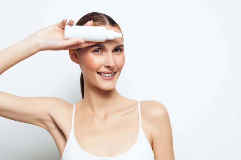 Smiling woman with clear skin holding white cosmetic bottle near eye on white background. Concept of skincare, beauty routine, health, wellness, and natural care with bright studio lighting.