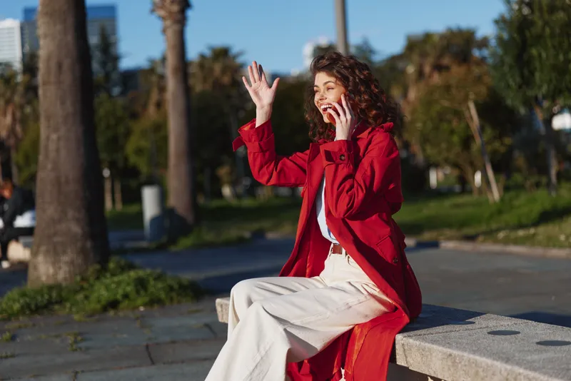 Young woman in red coat sitting on bench outdoors waving hand while talking on mobile phone sunny day casual outfit urban park background