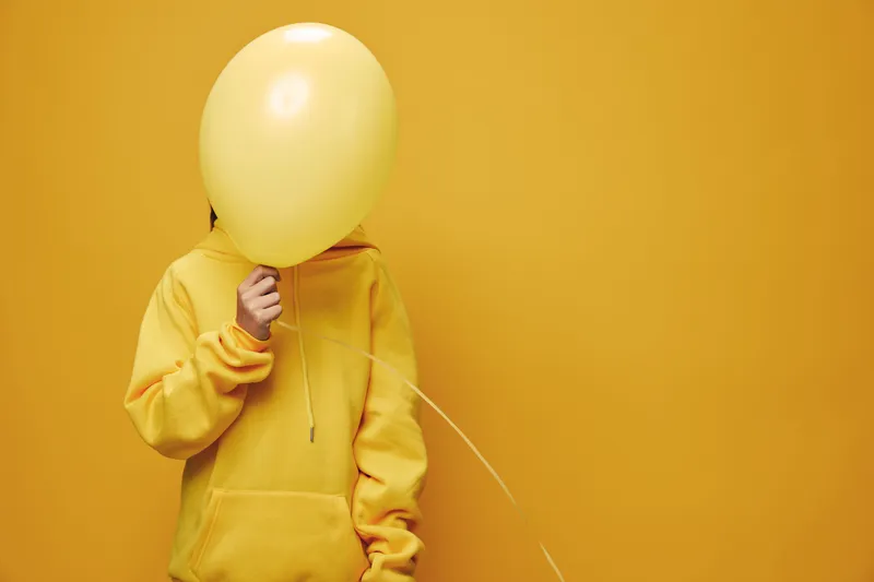 Person in yellow hoodie holding a yellow balloon covering the face standing against yellow background. Casual outfit and monochrome style with soft lighting in studio.