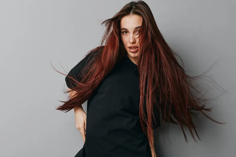 Young woman with long red hair wearing black sweater posing against gray background with dynamic hair movement and confident expression in studio portrait photography session.