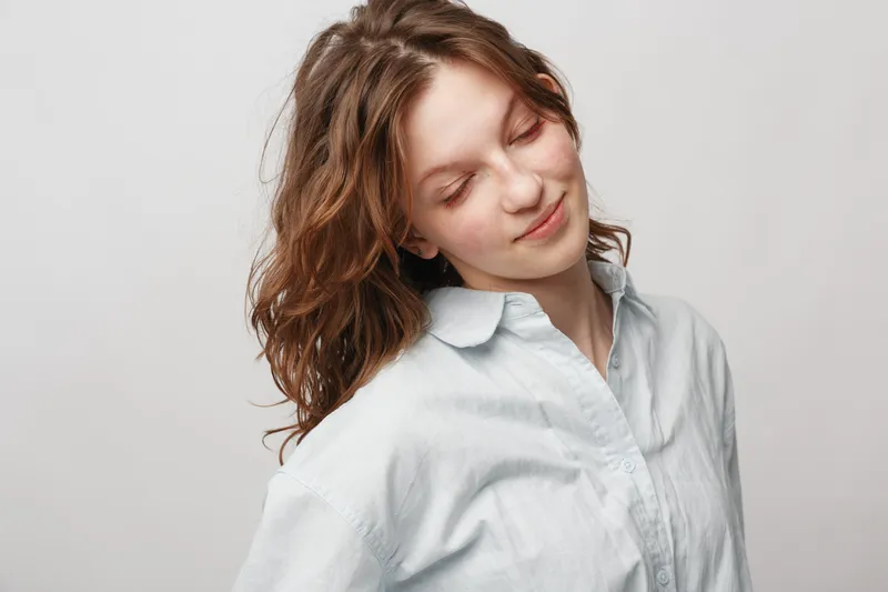 Young woman with wavy hair wearing white shirt looks down with gentle smile on light background. Concept of natural beauty, calmness, and soft emotions in portrait photography.