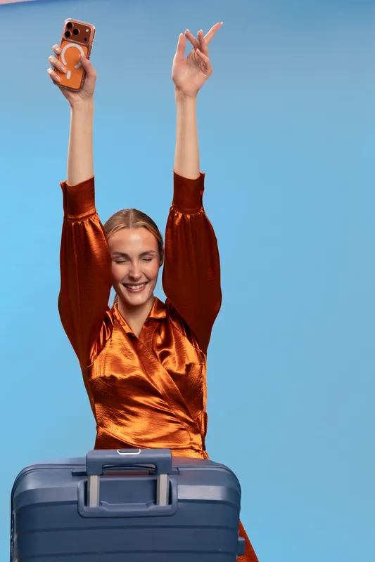 Happy young woman in orange satin dress raising arms with smartphone, standing behind blue suitcase. Concept for travel excitement, vacation, and joyful holiday moments.