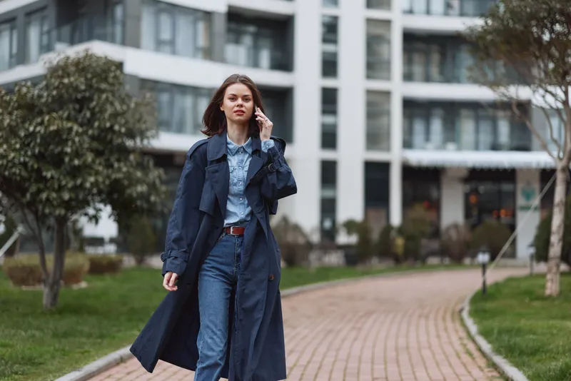 A stylish woman walks along a city sidewalk in a long trench coat and denim, talking on a smartphone. A candid urban scene showing confidence, fashion, and daily life.