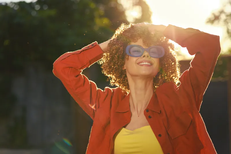 Smiling woman with curly hair wearing a red jacket and yellow top stands outdoors in warm sunshine, posing with sunglasses. Concept of casual lifestyle, confidence, and outdoor positivity