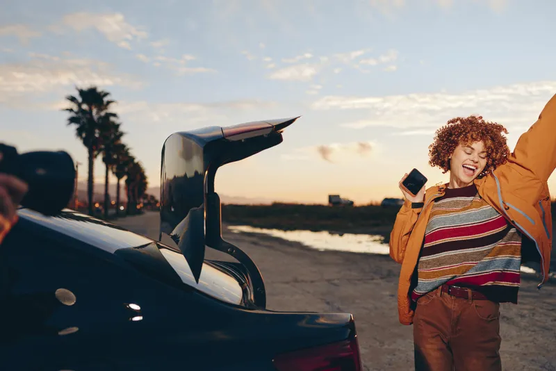 Woman lifestyle travel at sunset on beach, cheerful woman near car open trunk holds smartphone, casual orange jacket and striped shirt, laughing enjoying trip and smiling natural light