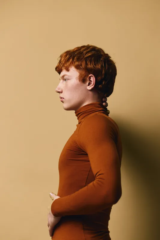 Profile of young man with red hair wearing brown turtleneck posing against beige background. Side view of thoughtful male model with natural skin and modern hairstyle in casual fashion.