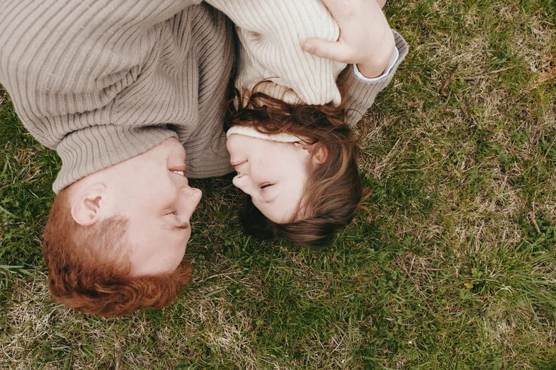 Two people lie on grass outdoors, sharing affection and warmth in a tender moment. A cozy lifestyle portrait captures togetherness, smile, and natural connection on a sunlit, calm day.