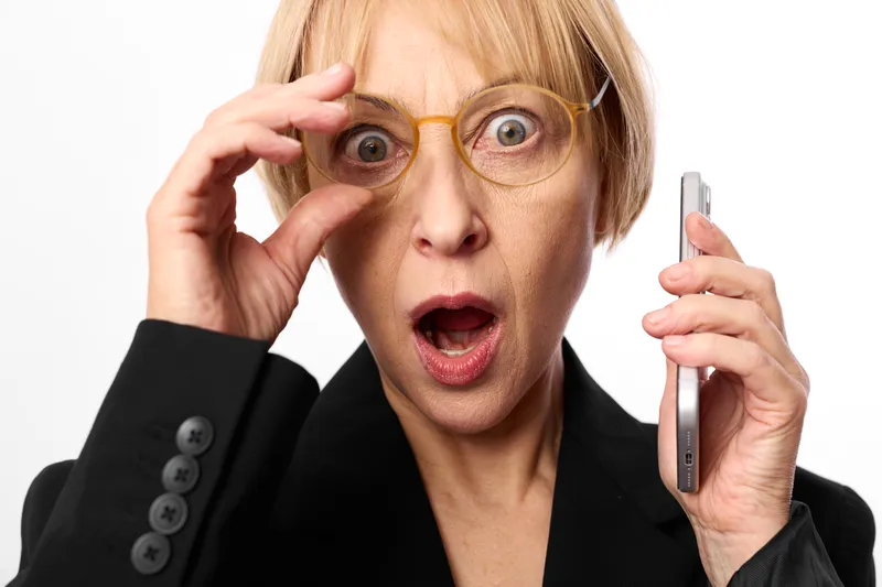 Businesswoman shocked and surprised, wearing glasses, holding phone to ear, tense stress panic expression, hand adjusting frames, white business background