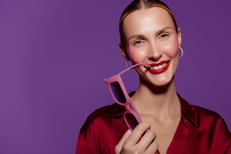 Smiling woman with red lipstick holds pink sunglasses near mouth on purple background. Concept of fashion, beauty, style, and confident female portrait with vibrant colors.